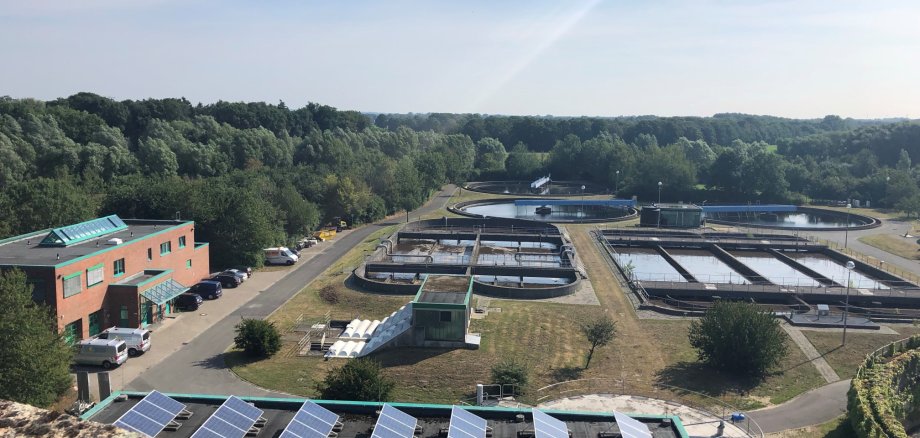 Ausblick über die gesamte Anlage des Abwasserwerks von oben. Mehrere Klärbecken und Verwaltunsgebäude links im Bild sichtbar. Strahlend blauer Himmel.