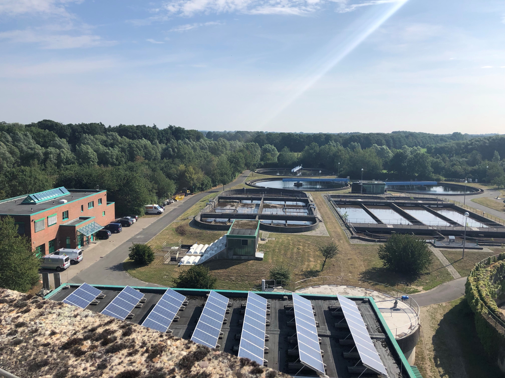 Ausblick über die gesamte Anlage des Abwasserwerks von oben. Mehrere Klärbecken und Verwaltunsgebäude links im Bild sichtbar. Strahlend blauer Himmel.