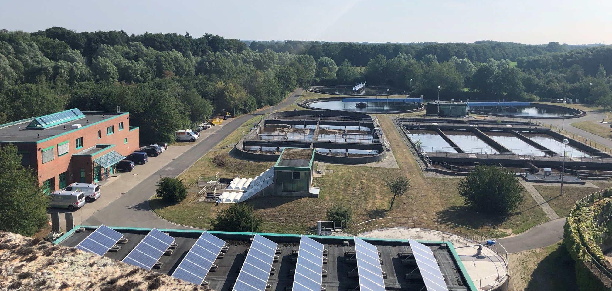 Ausblick über die gesamte Anlage des Abwasserwerks von oben. Mehrere Klärbecken und Verwaltunsgebäude links im Bild sichtbar. Strahlend blauer Himmel.