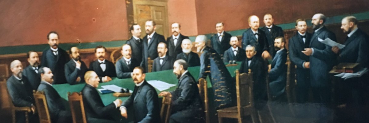 Ein gezeichnetes Bild von vielen Männern in schwarzen Anzügen aus vergangener Zeit. Es zeigt den Stadtrat, wie er damals ausgesehen hat. Die Männer sind teils sitzend, teils stehend um einen Tisch versammelt, auf dem zwei Dokumente liegen. Einige Männer unterhalten sich angeregt miteinander.