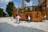 Mehrere junge Kinder im Alter von ca. 5 Jahren rennen auf ein Piratenschiff aus Holz zu, das zu einem Spielgerät umgebaut wurde. Das Piratenschiff ist weitaus größer als die Kinder.