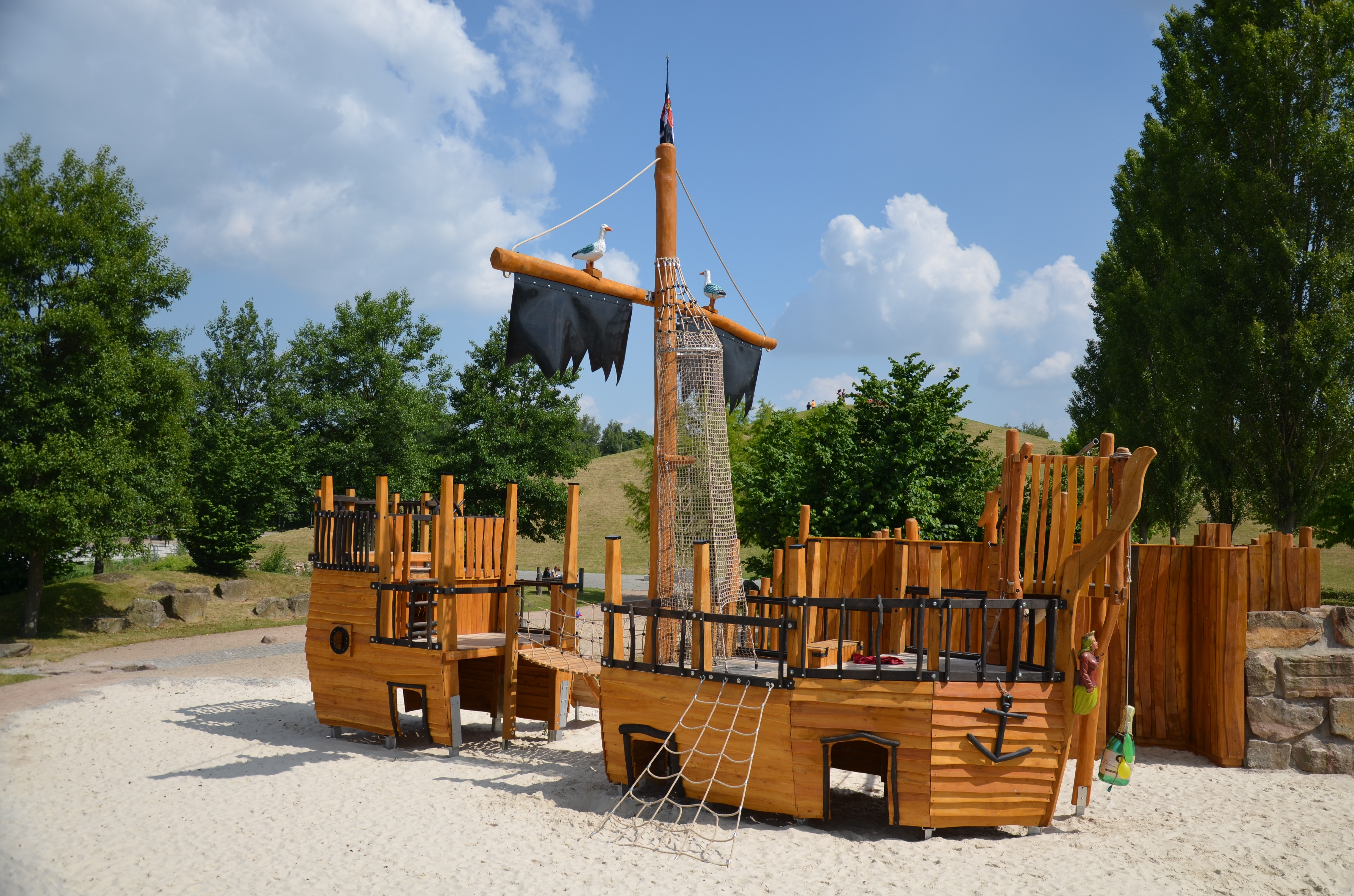 Ein Piratenschiff aus Holz, das zu einem übergroßen Spielgerät umfunktioniert wurde. Strahlend blauer Himmel, im Hintergrund sieht man Begrünung.