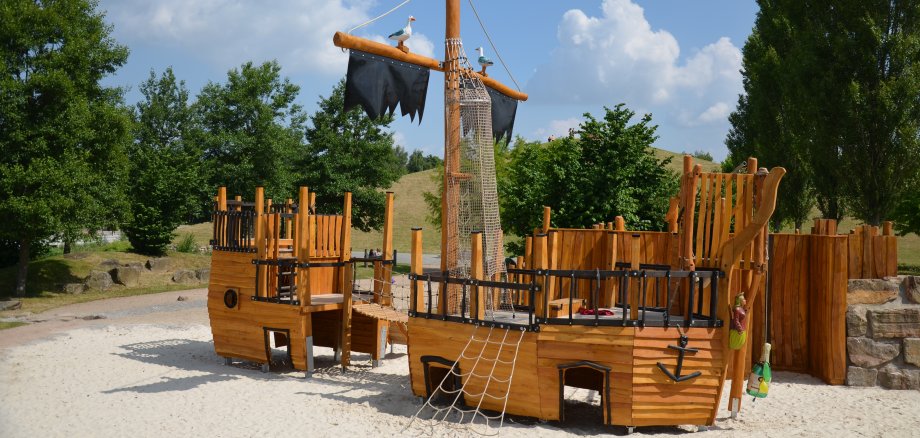 Ein Piratenschiff aus Holz, das zu einem übergroßen Spielgerät umfunktioniert wurde. Strahlend blauer Himmel, im Hintergrund sieht man Begrünung.