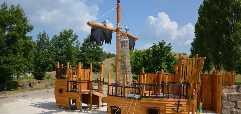 Ein Piratenschiff aus Holz, das zu einem übergroßen Spielgerät umfunktioniert wurde. Strahlend blauer Himmel, im Hintergrund sieht man Begrünung.