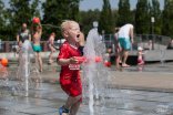 Ein kleiner blonder Junge mit rotem T-Shirt und roter Sporthose rennt spielend durch das Wasserlabyrinth, aus dem eine Fontäne heraus schießt. Im Hintergrund sind verschwommen weitere Personen unterschiedlichen Alters in Badekleidung zu sehen. Ein kleiner blonder Junge mit rotem T-Shirt und roter Sporthose rennt spielend durch das Wasserlabyrinth, aus dem eine Fontäne heraus schießt. Im Hintergrund sind verschwommen weitere Personen unterschiedlichen Alters in Badekleidung zu sehen.