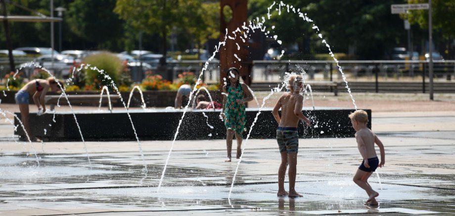 Drei Kinder von hinten die unter mehren Fontänen des Wasserlabyrinths umher springen.