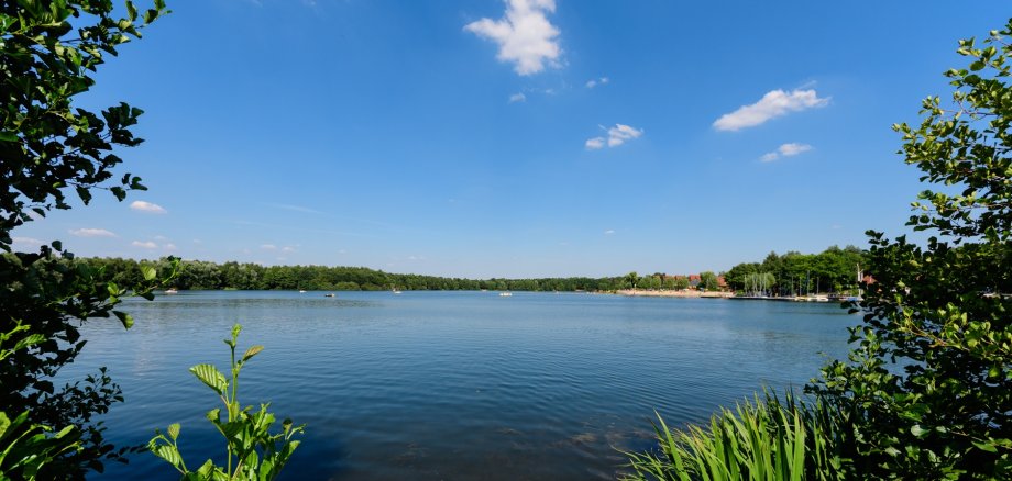 Der See in seiner ganzen Größe mit blauen Himmel und angedeutetem grünen Gestrüpp um den See herum.