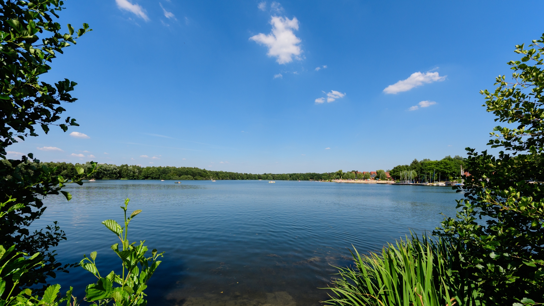 Der See in seiner ganzen Größe mit blauen Himmel und angedeutetem grünen Gestrüpp um den See herum.