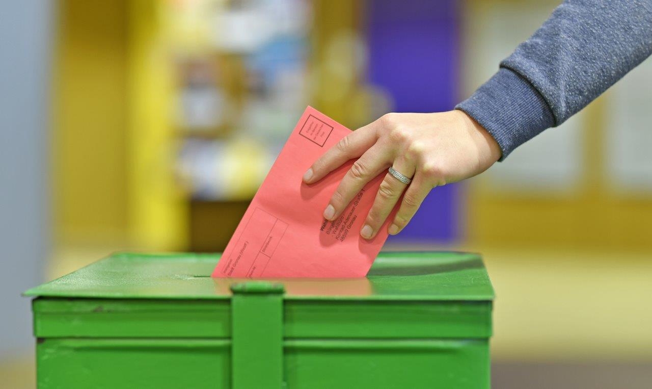 Eine Hand wirft einen roten Umschlag in eine grüne Wahlurne.