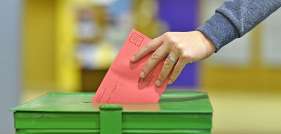 Eine Hand wirft einen roten Umschlag in eine grüne Wahlurne.