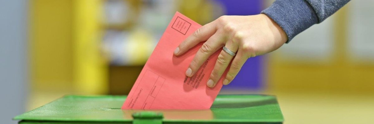 Eine Hand wirft einen roten Umschlag in eine grüne Wahlurne. Eine Hand wirft einen roten Umschlag in eine grüne Wahlurne.