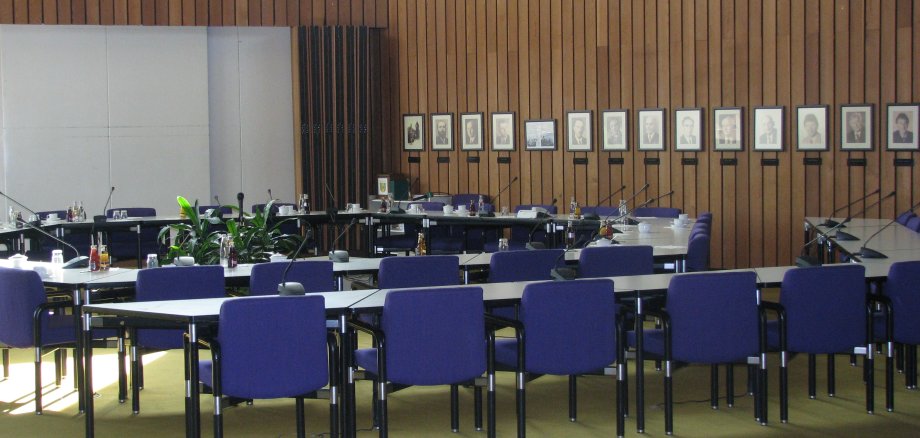 Der Sitzungssaal mit Tischen, Stühlen und einer Leinwand.