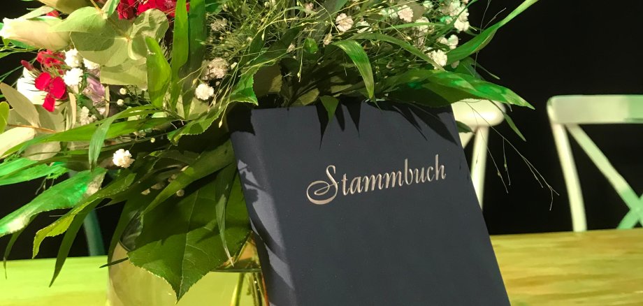 Ein buntes Blumenbouquet in einer Glasvase vor dem ein schwarzes Stammbuch mit weißer Aufschrift steht.