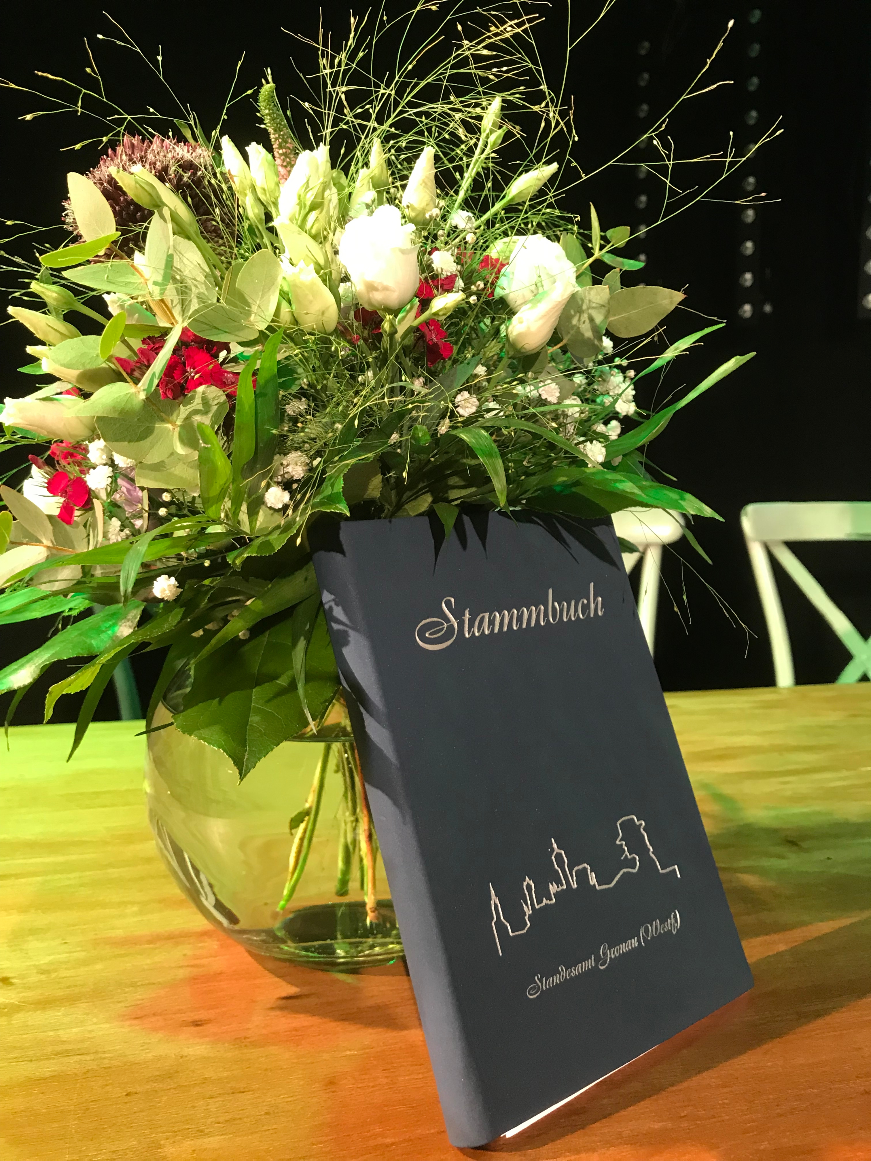 Ein buntes Blumenbouquet in einer Glasvase vor dem ein schwarzes Stammbuch mit weißer Aufschrift steht.