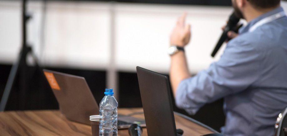 Eine Person sitzt vor zwei Laptops an einem Tisch und hält ein Mikrofon in der Hand.