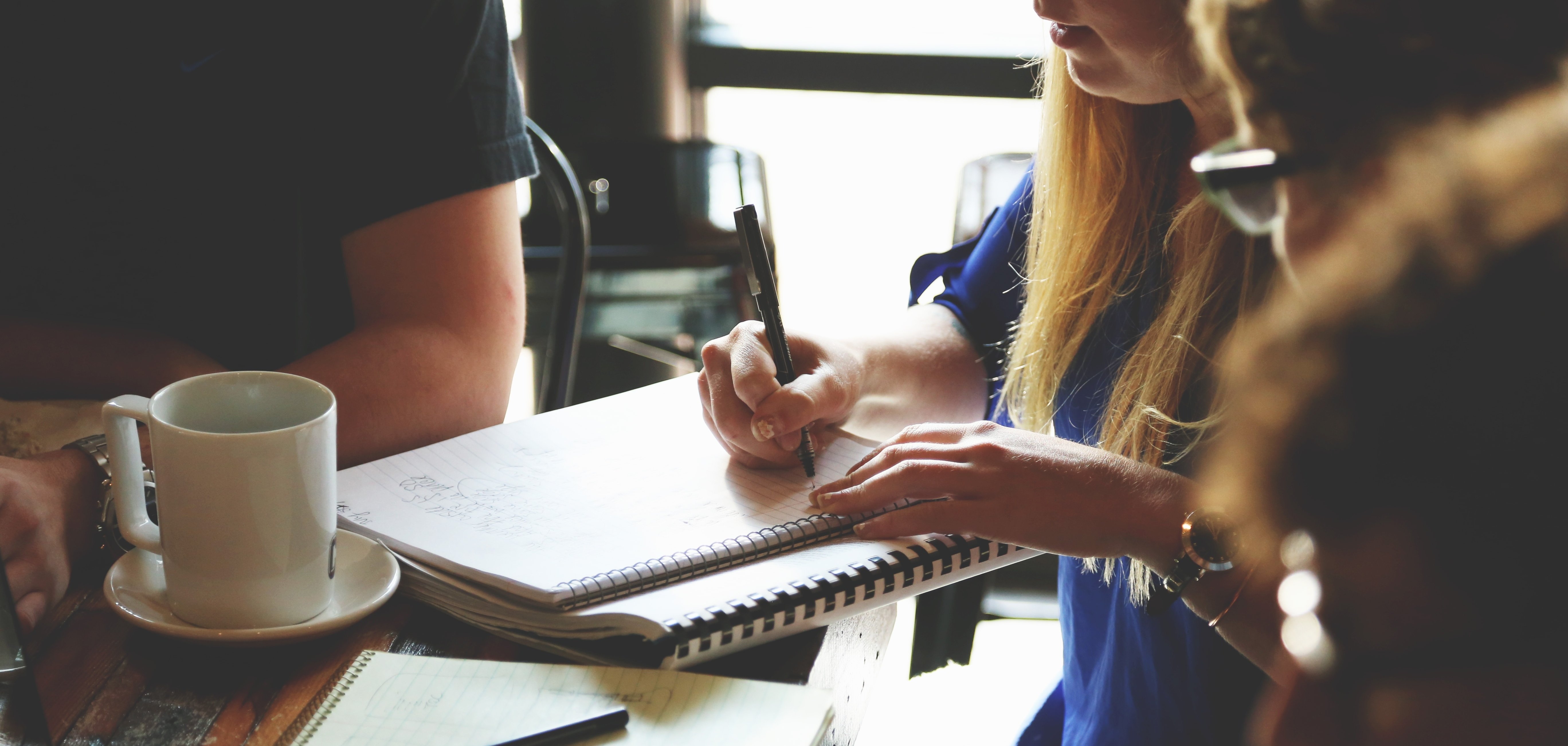 Drei Personen sitzen um einen Tisch, auf dem ein Laptop, mehrere Stifte und eine Kaffeetasse stehen. Der Fokus des Bildes liegt auf dem Tisch, die Personen sind nicht erkennbar. Eine der Personen schreibt etwas auf einen Collegeblock.