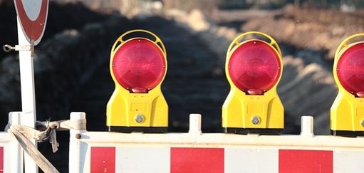 Man sieht eine Baustellenabsperrung mit roten Lichtern vor einer Straße.