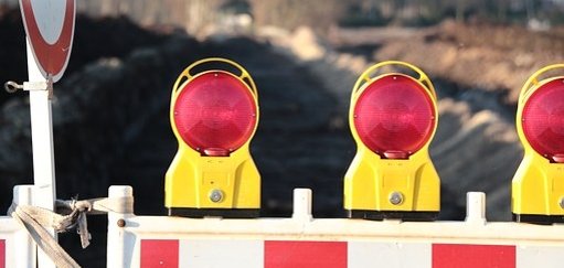 Man sieht eine Baustellenabsperrung mit roten Lichtern vor einer Straße.