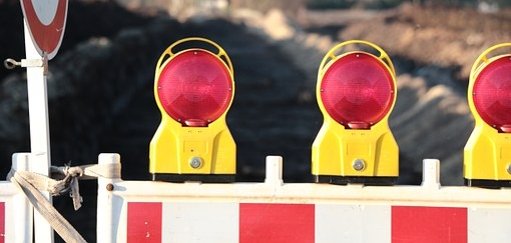 Man sieht eine Baustellenabsperrung mit roten Lichtern vor einer Straße.