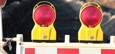 Man sieht eine Baustellenabsperrung mit roten Lichtern vor einer Straße.