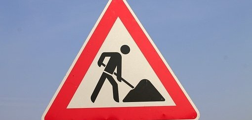 Man sieht ein rotweißes Baustellenschild mit einem schwarzen Männchen mit Schüppe und Sandhaufen.