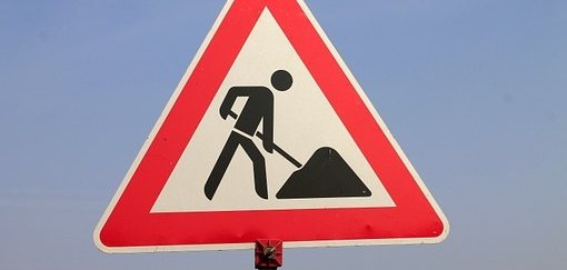 Man sieht ein rotweißes Baustellenschild mit einem schwarzen Männchen mit Schüppe und Sandhaufen.