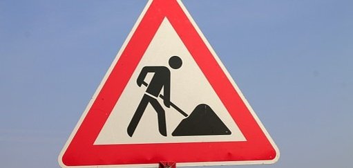 Man sieht ein rotweißes Baustellenschild mit einem schwarzen Männchen mit Schüppe und Sandhaufen.