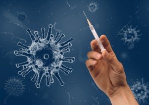Impfen: Spritze Eine Hand hält eine Spritze hoch, im Hintergrund ist das Coronavirus.