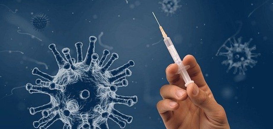 Eine Hand hält eine Spritze hoch, im Hintergrund ist das Coronavirus.