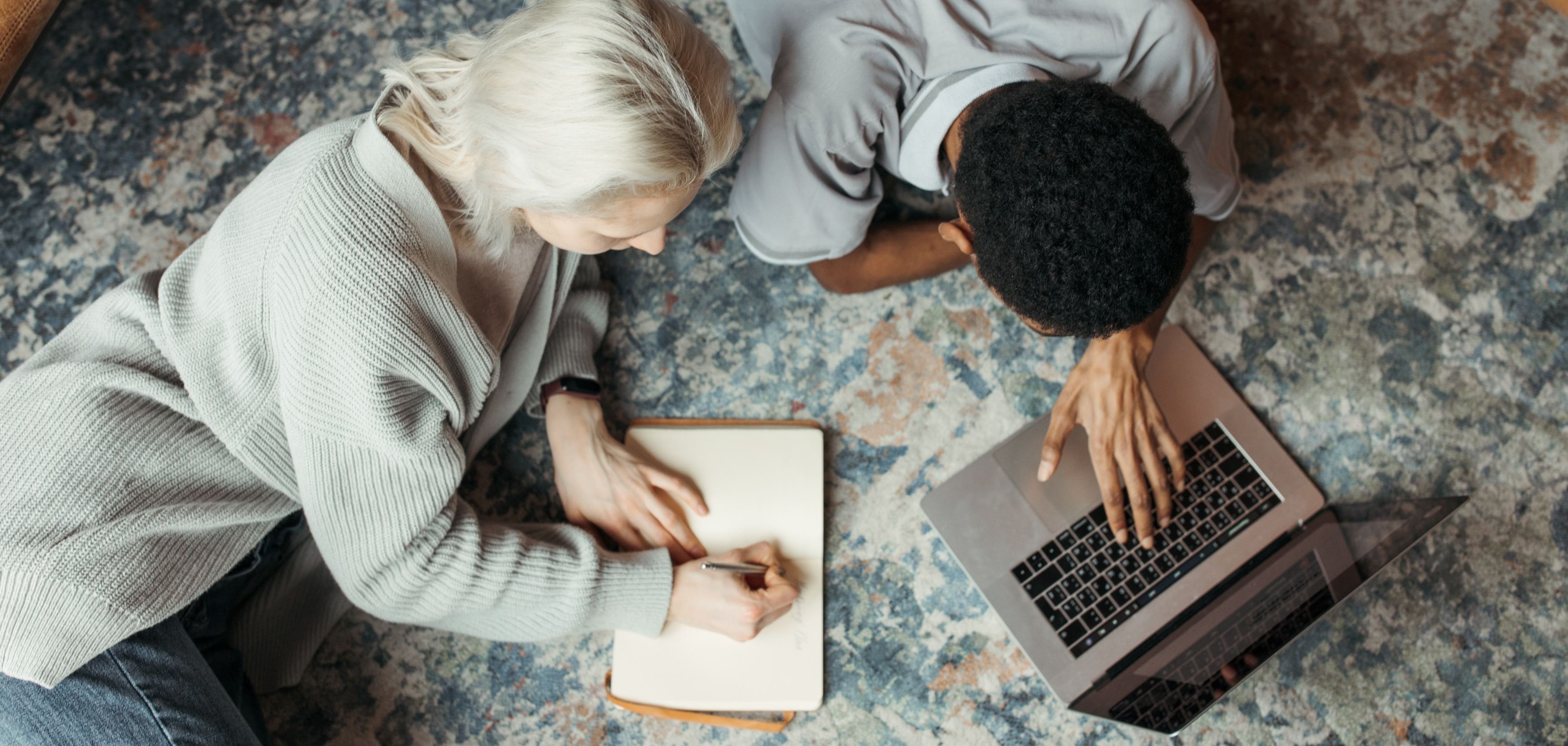 Zwei Personen liegen auf dem Boden und schreiben Texte in ein Buch und einen Laptop.