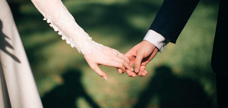 Man sieht den Arm einer Braut und den Arm eines Bräutigams. Die beiden strecken ihren Arm zueinander und halten im Ansatz Händchen.