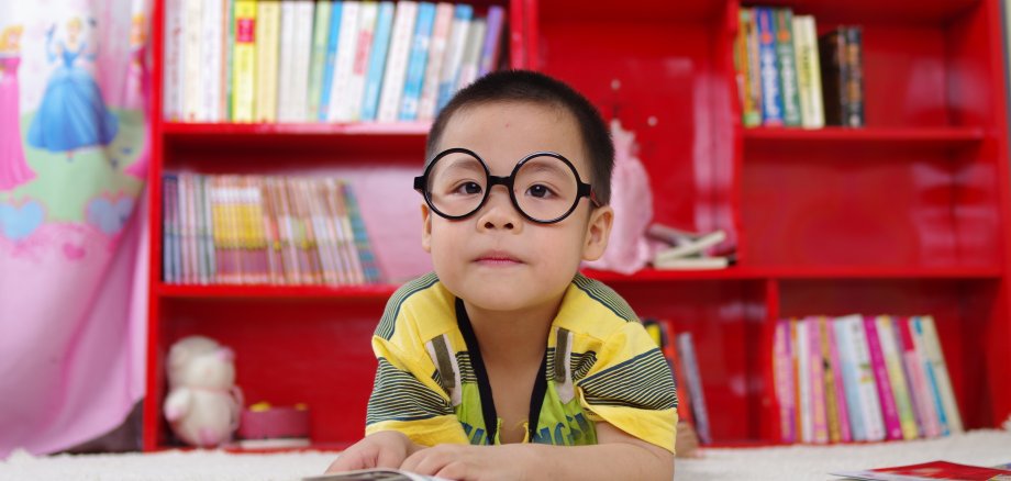 Ein kleines Kind mit schwarzer Brille sitzt vor einem rotem Bücherregal.