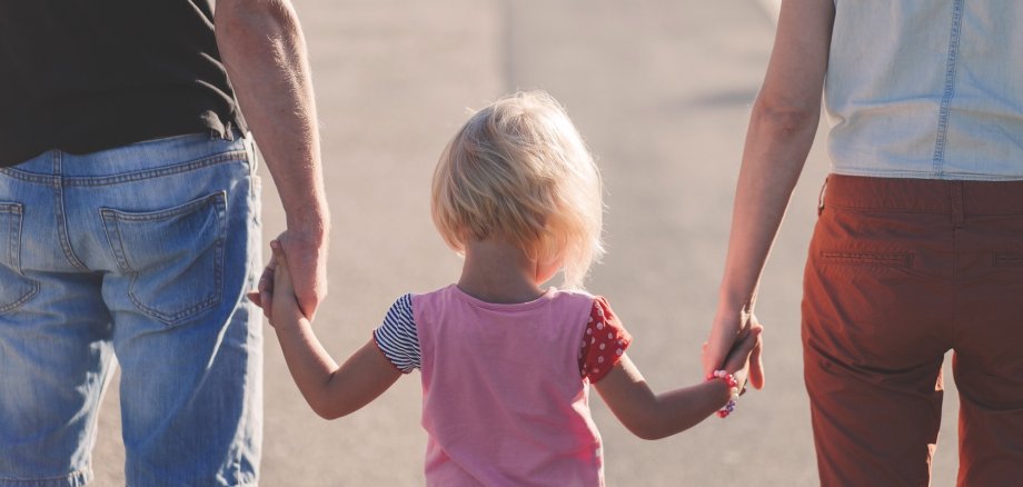 Eine Frau und ein Mann, von denen man nur die Hälfte des Rückens ab Schulterhöhe sieht, halten mit jeweils einer Hand die Hände eines kleinen blonden Mädchens, das auch nur von hinten zu sehen ist und in der Mitte zwischen dem Mann und der Frau läuft.