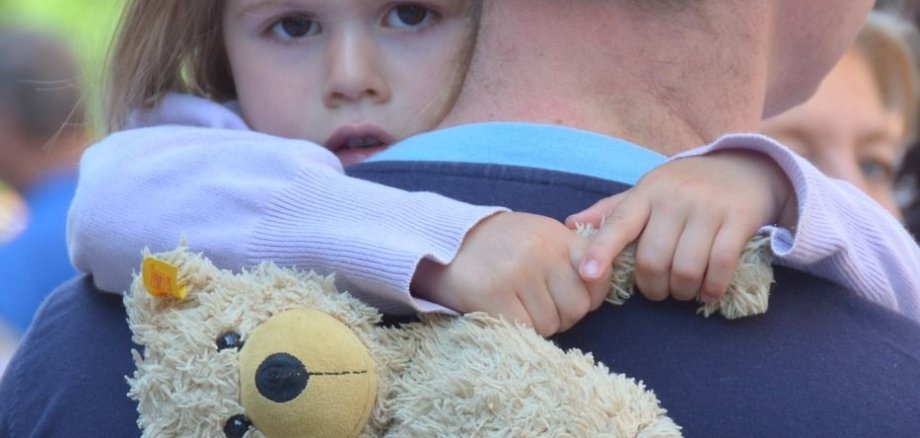 Ein Kind ist auf dem Arm seines Vaters und hält einen Teddy in der Hand.