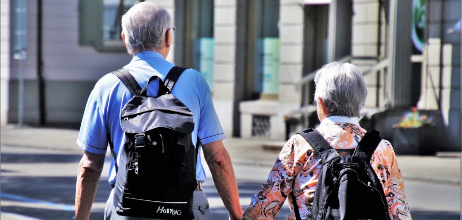 Es ist ein älteres Ehepaar dargestellt die jeweils einen Rucksack aufhaben und spazieren