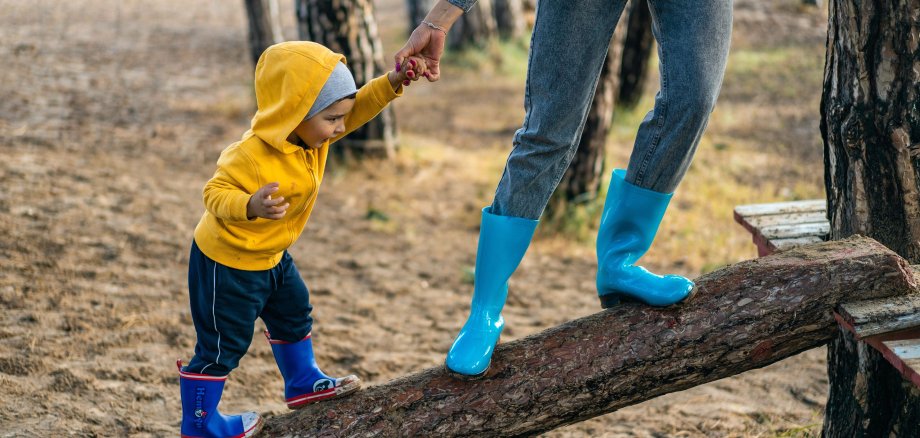 Ein Kind läuft mit seiner Mutter an der Hand auf einem Baumstamm.