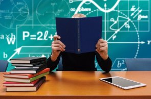 Schule Eine Lehrerein sitzt mit einem Buch in der Hand vor einer Tafel.