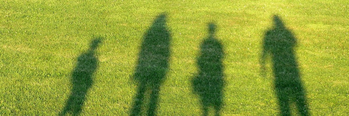 Vier Schatten von Menschen unterschiedlicher Größe, Geschlecht und Alter auf einer Rasenfläche.