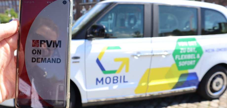 Eine Hand hält ein Handy, auf dem die App RVM On-Demand geöffnet ist. Dahinter ist das G-Mobil zu sehen.