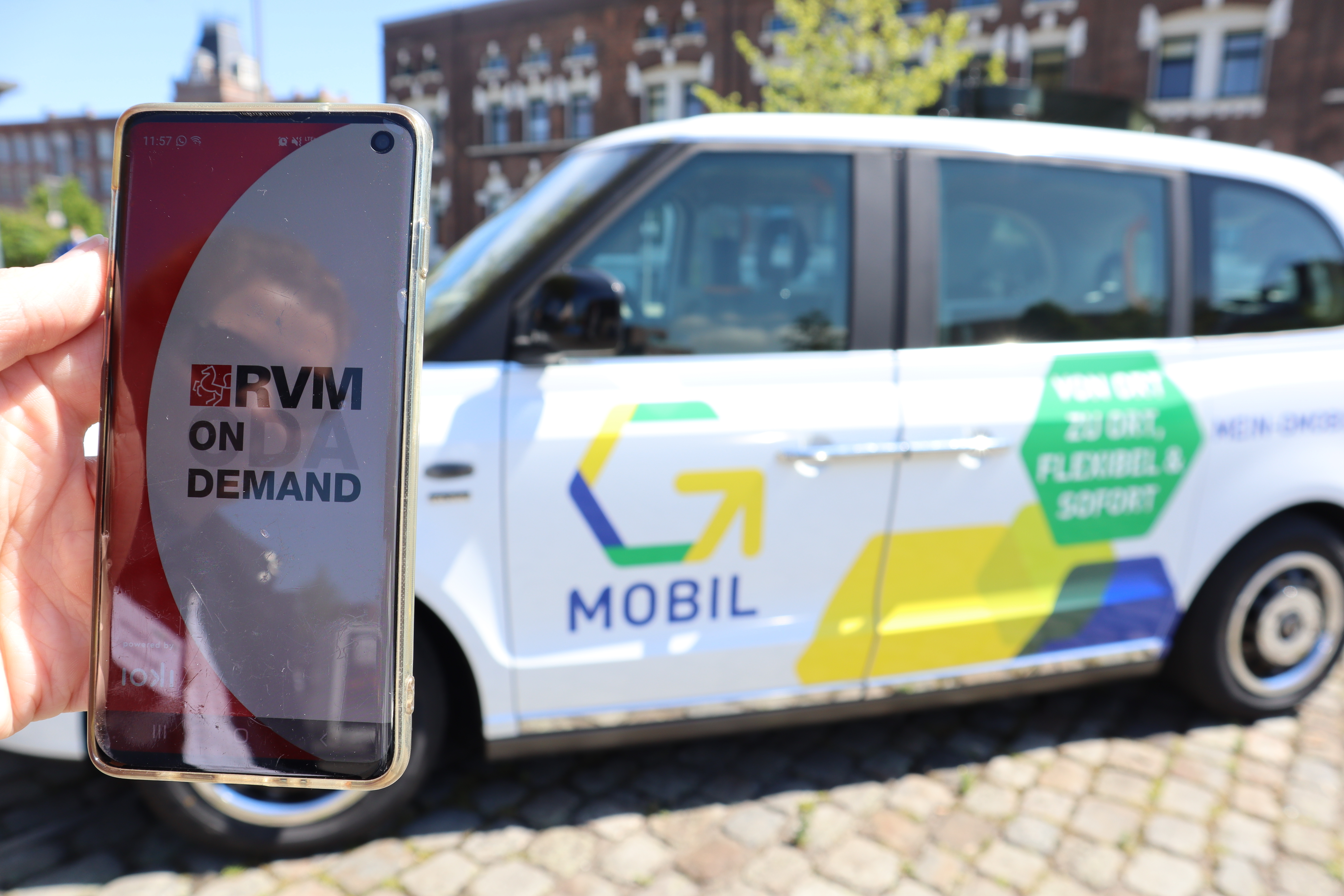 Eine Hand hält ein Handy, auf dem die App RVM On-Demand geöffnet ist. Dahinter ist das G-Mobil zu sehen.