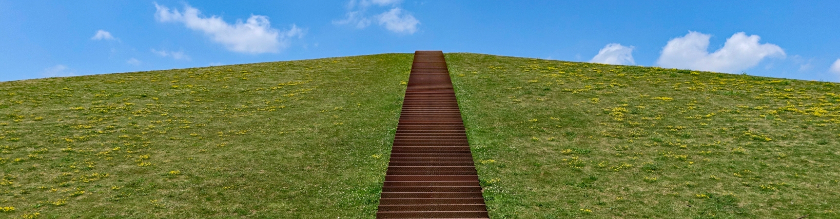 Treppenaufstieg zum grün bepflanzten LAGA Berg von unten, im Hintergrud ein strahlend blauer Himmel.