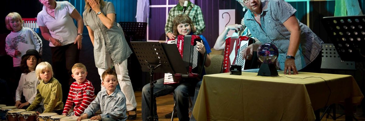 Eine Gruppe gemischten Alters und Geschlechts schaut erstaunt nach rechts. Einige der Personen halten ein Instrument in der Hand. Es handelt sich um eine Szene aus einer Aufführung der Musikschule.
