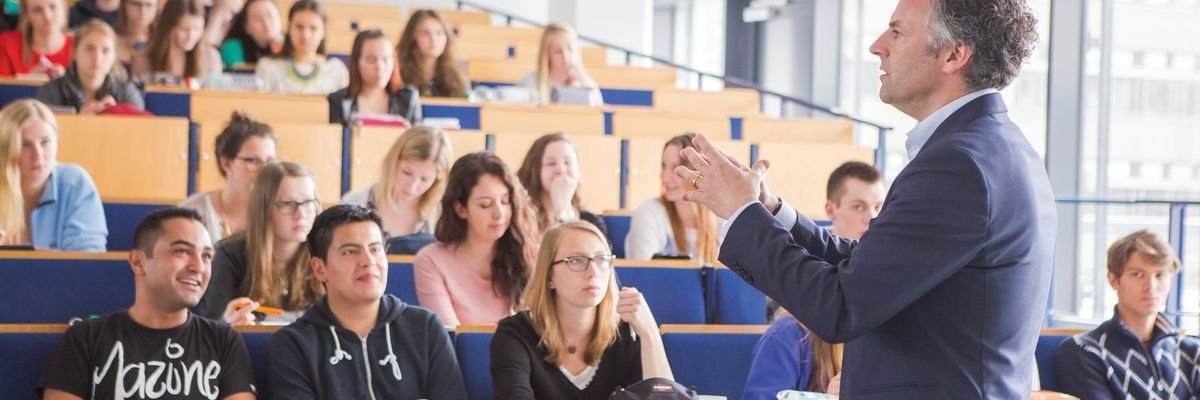 Auf dem Foto sieht man einen Dozenten in einem mittelgroßen Hörsaal. Er spricht zu seinen Studenten und gestikuliert dabei mit den Händen. Die Studenten sind unterschiedlichen Alters, Geschlechts und Nationalität.