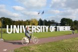 Auf einer Wiese stehen große weiße Buchstaben, die "University of Twente" buchstabieren. Vor den Buchstaben flitzt eine Radfahrerin durch das Bild.