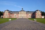Auf dem Foto ist das Schloss der Universität Münster zu sehen, mit dem Blick aus der Richtung Innenstadt. Die Front ist beige/rotbraun, mit einem dunklen Dach. Der Himmel ist strahlend blau.