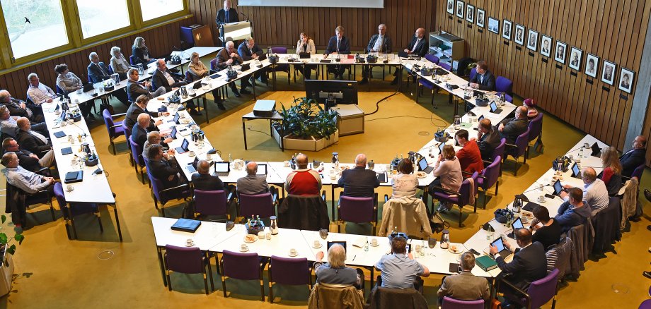 Vogelperspektive auf den Sitzungssaal der Stadt Gronau, in dem die Ratsmitglieder an ihren Tischen sitzen und einem Vortrag von einem weiteren Ratsmitglied lauschen.