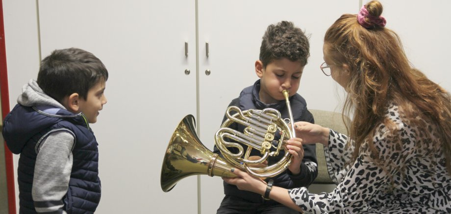 zu sehen sind zwei Kinder und eine Erwachsene Person. Die Erwachsene Person zeigt einem Kind ein Musikinstrument.