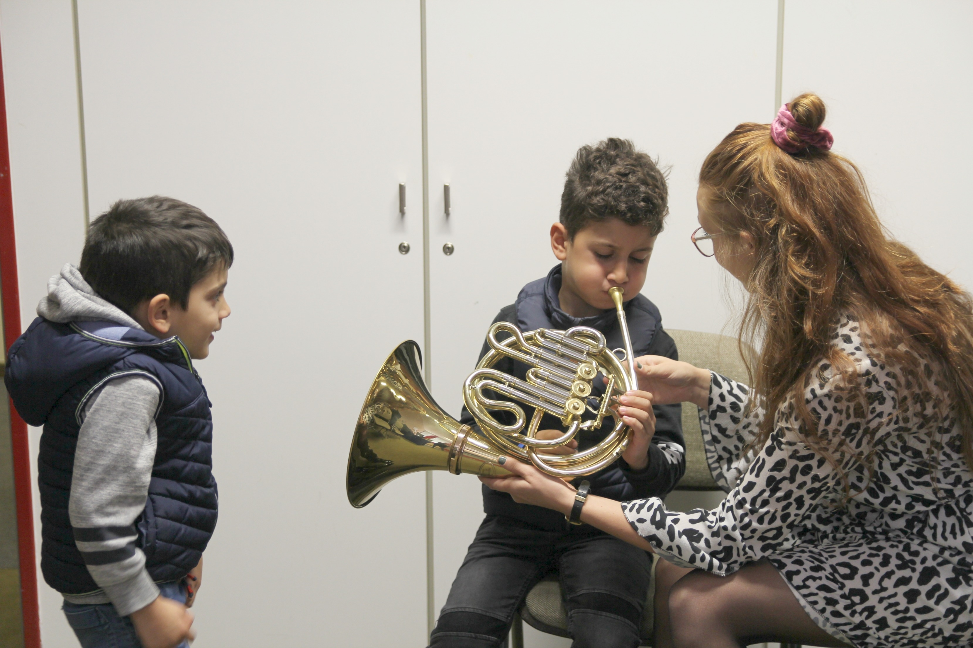 zu sehen sind zwei Kinder und eine Erwachsene Person. Die Erwachsene Person zeigt einem Kind ein Musikinstrument.