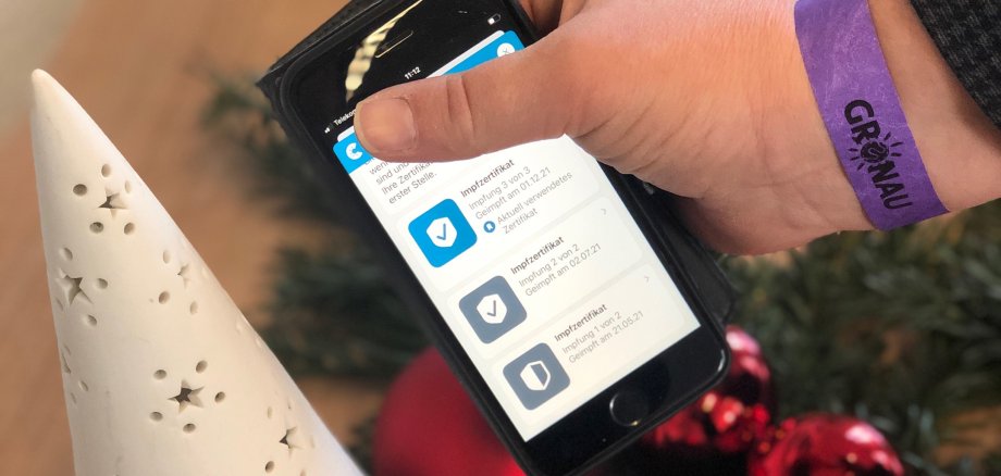 Eine Hand mit einem Bändchen um das Handgelenk hält ein Handy, auf dem der Impfnachweis zu sehen ist.