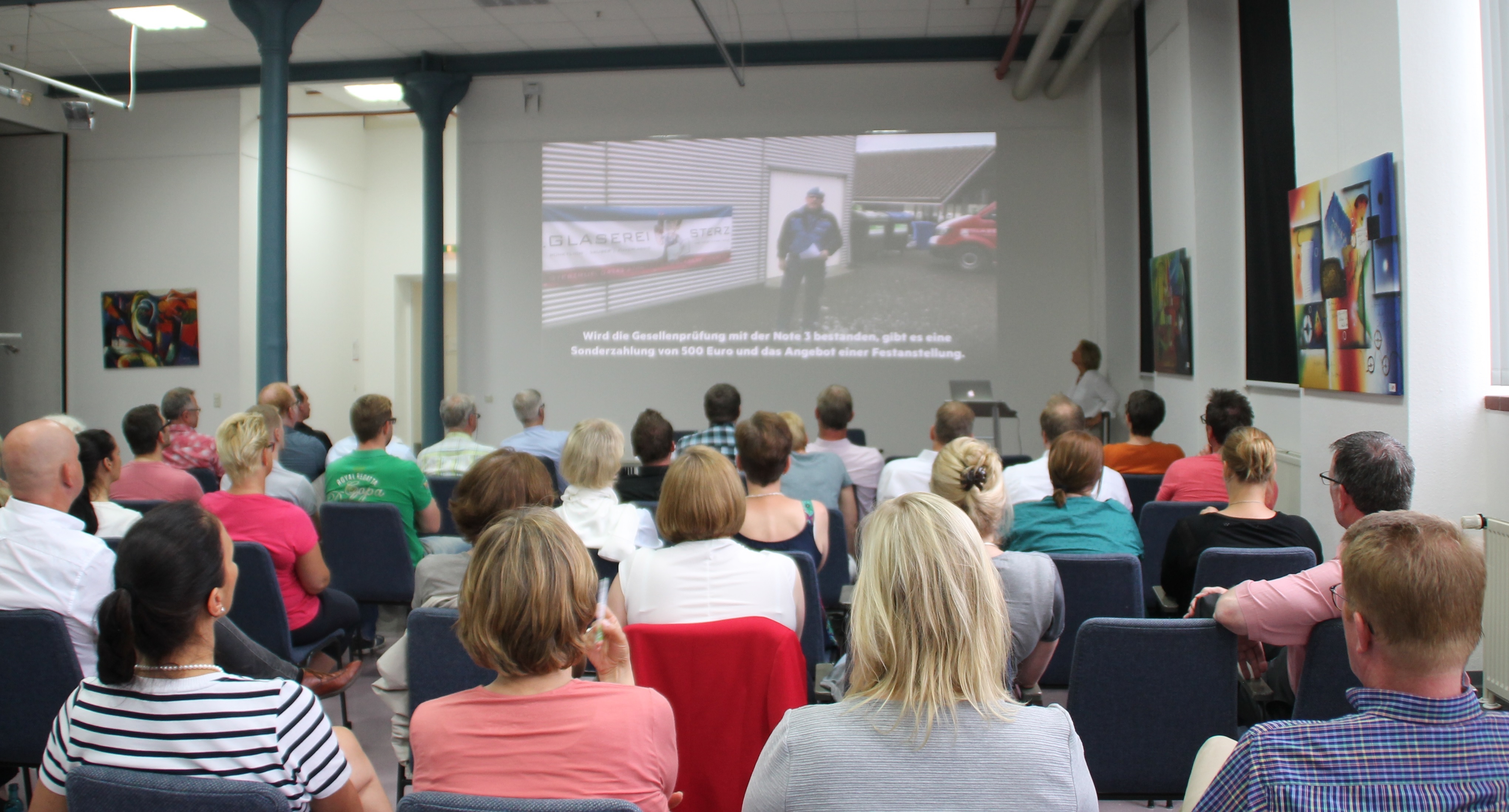 Ein Publikum sitzt auf Stühlen vor einer Bühne mit Leinwand.
